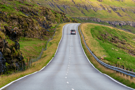 View of a car driving on a winding road through the green rocky hills of the Faroe Islands. View of a car driving on a winding road through the green rocky hills of the Faroe Islands.