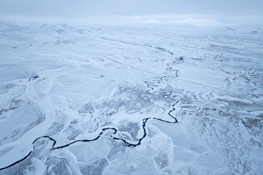 Aerial view of a winding river flowing through a vast snow covered landscape with distant mountains under a cloudy sky in Egilsstadir Mulathing Iceland. Aerial view of a winding river flowing through a vast snow covered landscape with distant mountains under a cloudy sky in Egilsstadir Mulathing Iceland.
