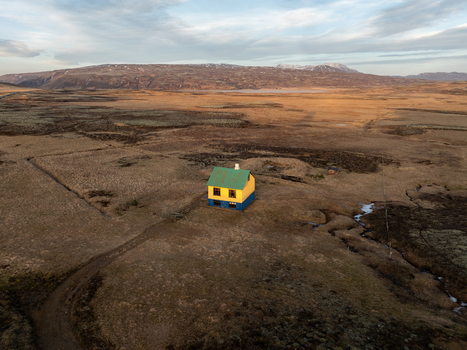 Aerial view of a small yellow and blue house standing alone in a vast rugged landscape with rolling hills and a winding stream in Reykjanesbaer Iceland. Aerial view of a small yellow and blue house standing alone in a vast rugged landscape with rolling hills and a winding stream in Reykjanesbaer Iceland.