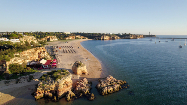 Aerial view of the sandy beach at Ferragudo featuring rocky cliffs a coastal restaurant and sailboats anchored in the calm blue water in Ferragudo Faro District Portugal.