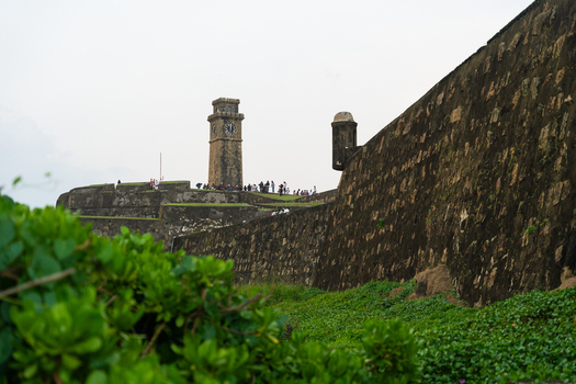 View of the Galle Fort Clock Tower and historic stone ramparts with people walking along the fortifications under an overcast sky in Galle Southern Province Sri Lanka.