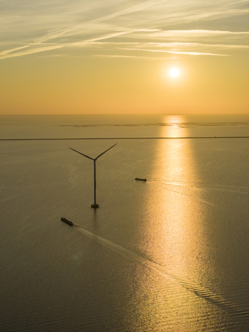 Aerial view of an offshore wind turbine and cargo ships navigating the calm sea during a golden sunset with a distant dike Netherlands. Aerial view of an offshore wind turbine and cargo ships navigating the calm sea during a golden sunset with a distant dike Netherlands.