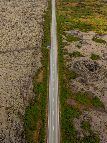 Aerial view of a straight asphalt road cutting through a vast volcanic lava field with mossy green patches and dark rocky terrain under an overcast sky. Aerial view of a straight asphalt road cutting through a vast volcanic lava field with mossy green patches and dark rocky terrain under an overcast sky.