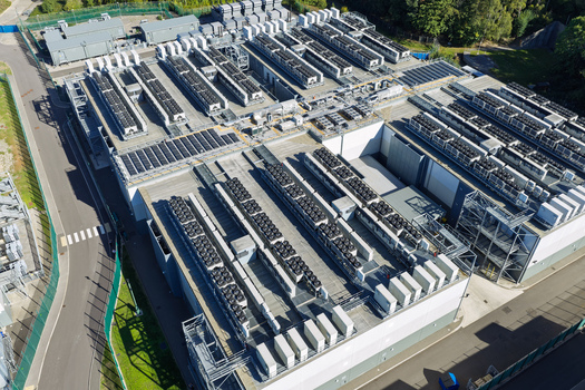 Aerial view of a large data center facility featuring rows of industrial cooling units on the roof surrounded by secure fencing and paved roads in the United Kingdom.