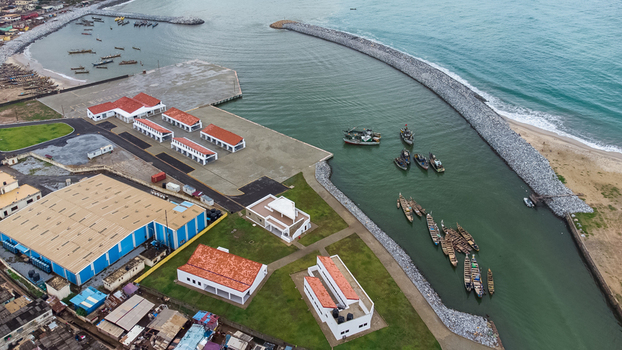 Aerial view of the Elmina harbor showing the breakwater fishing boats docked in the calm water and nearby industrial buildings with red tiled roofs in Elmina Central Region Ghana. Aerial view of the Elmina harbor showing the breakwater fishing boats docked in the calm water and nearby industrial buildings with red tiled roofs in Elmina Central Region Ghana.