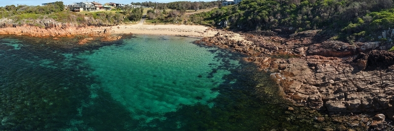 Aerial view of clear turquoise waters meeting rugged reddish cliffs and a sandy beach beneath coastal homes Actual City Name New South Wales Australia. Aerial view of clear turquoise waters meeting rugged reddish cliffs and a sandy beach beneath coastal homes Actual City Name New South Wales Australia.