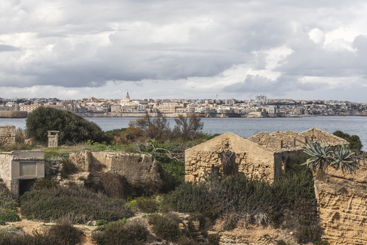 View of the historic city skyline across the water from ancient stone ruins and green vegetation under a cloudy sky Syracuse Sicily Italy.
