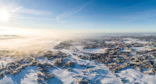 Aerial view of a vast snow covered landscape featuring frost laden forests open fields and a layer of morning mist under a bright sun in Latvia. Aerial view of a vast snow covered landscape featuring frost laden forests open fields and a layer of morning mist under a bright sun in Latvia.