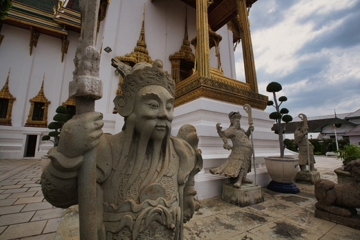 View of stone guardian statues at The Grand Palace with ornate golden architecture and paved courtyard in Bangkok Thailand. View of stone guardian statues at The Grand Palace with ornate golden architecture and paved courtyard in Bangkok Thailand.