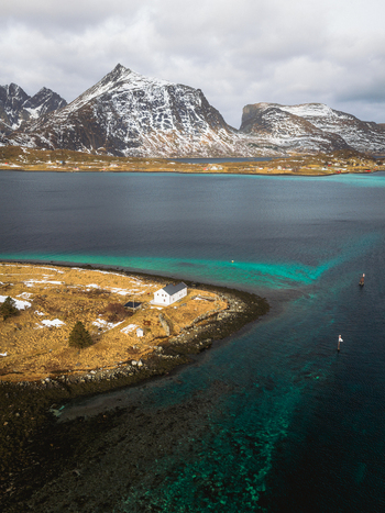 Aerial view of turquoise waters meet rugged shores near Kåkernbrua with snow dusted mountains looming under a cloudy sky Ramberg Nordland Norway. Aerial view of turquoise waters meet rugged shores near Kåkernbrua with snow dusted mountains looming under a cloudy sky Ramberg Nordland Norway.