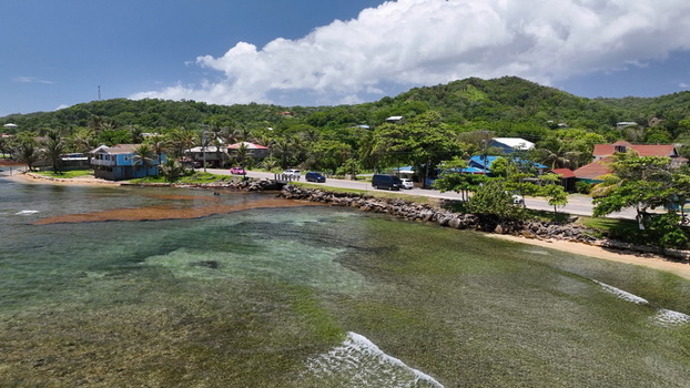 Aerial view of the coastal landscape featuring clear turquoise waters a rocky shoreline a road with passing vehicles and lush green hills in West End Bay Islands Department Honduras. Aerial view of the coastal landscape featuring clear turquoise waters a rocky shoreline a road with passing vehicles and lush green hills in West End Bay Islands Department Honduras.