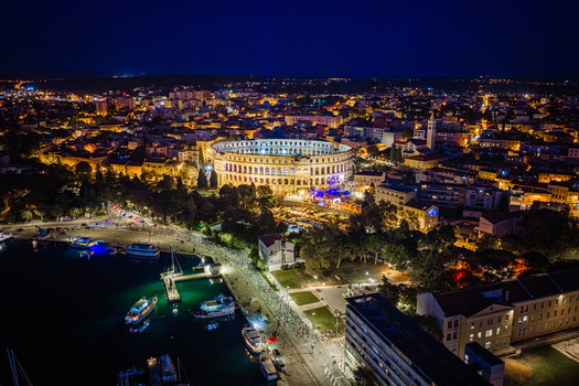 Aerial view of the illuminated Pula Arena a historic Roman amphitheater set against the vibrant city lights and harbor of Pula Istria County Croatia. Aerial view of the illuminated Pula Arena a historic Roman amphitheater set against the vibrant city lights and harbor of Pula Istria County Croatia.