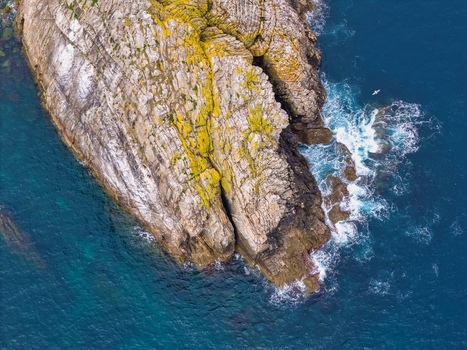 Aerial view of the rugged cliffs and mossy rock formations at Silence Beach Viewpoint with crashing waves and a seagull over the deep blue sea in Cudillero Asturias Spain.