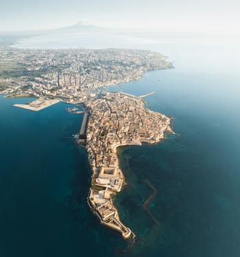 Aerial view of the Island of Ortigia and Maniace Castle surrounded by the blue Mediterranean sea with Mount Etna in the background in Syracuse Sicily Italy.