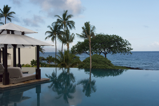 View of an infinity pool with lounge chairs and umbrellas overlooking the ocean with palm trees in the background in Maui County Hawaii United States.