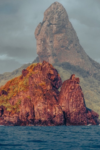 View of the iconic Morro do Pico rock formation rising above the blue ocean with reddish volcanic rocks in the foreground in Fernando de Noronha State of Pernambuco Brazil. View of the iconic Morro do Pico rock formation rising above the blue ocean with reddish volcanic rocks in the foreground in Fernando de Noronha State of Pernambuco Brazil.
