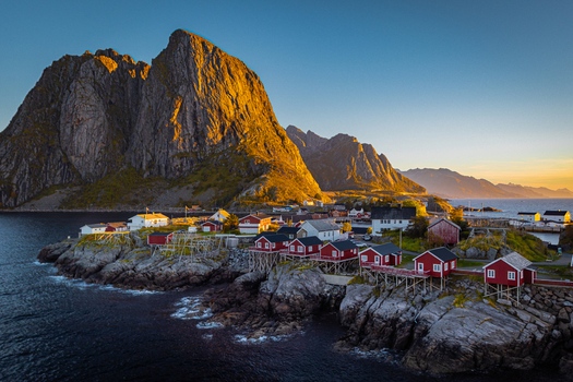 View of the iconic red rorbuer cabins on stilts in the fishing village of Hamnoy set against dramatic rocky mountains under golden hour light in Hamnoy Nordland Norway.