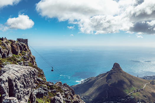 View of the Table Mountain cable car descending towards the Atlantic Ocean with Lion s Head peak under a cloudy blue sky in Cape Town Western Cape South Africa. View of the Table Mountain cable car descending towards the Atlantic Ocean with Lion s Head peak under a cloudy blue sky in Cape Town Western Cape South Africa.