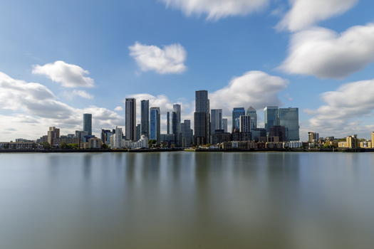 View of the Canary Wharf skyline with modern skyscrapers reflecting in the calm waters of the River Thames under a bright blue sky with scattered clouds in London England United Kingdom. View of the Canary Wharf skyline with modern skyscrapers reflecting in the calm waters of the River Thames under a bright blue sky with scattered clouds in London England United Kingdom.