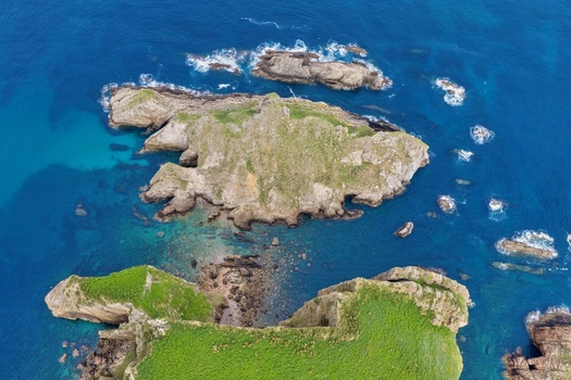 Aerial view of Silence Beach Viewpoint featuring rugged rocky islets and green cliffs surrounded by deep blue ocean water and crashing waves in Cudillero Asturias Spain.