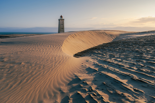 View of the historic Rubjerg Knude lighthouse standing amidst vast shifting sand dunes under a clear sky at sunset in Lokken Denmark. View of the historic Rubjerg Knude lighthouse standing amidst vast shifting sand dunes under a clear sky at sunset in Lokken Denmark.