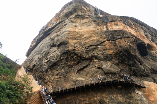 View of the massive Sigiriya Rock Fortress with tourists climbing the metal stairs along the ancient rock face Sigiriya Central Province Sri Lanka. View of the massive Sigiriya Rock Fortress with tourists climbing the metal stairs along the ancient rock face Sigiriya Central Province Sri Lanka.