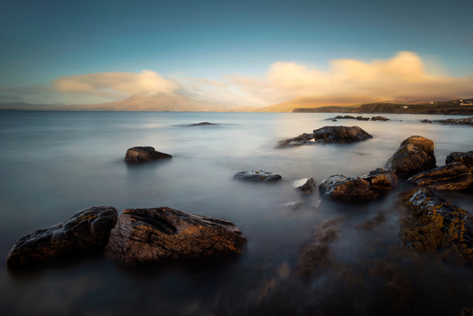 View of dark rocks in the calm sea with misty mountains under a soft golden sky at Lettergesh Beach County Galway Ireland.