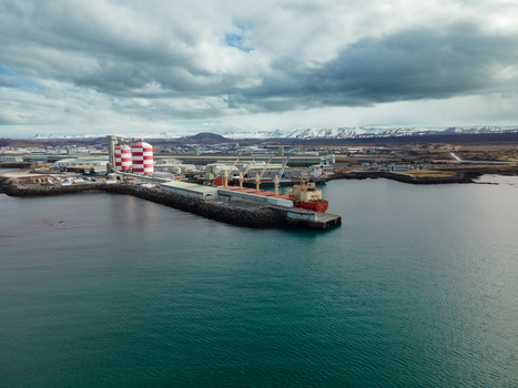Aerial view of an industrial port with a cargo ship docked near red and white striped silos and snow capped mountains Hafnarfjordur Hafnarfjardarkaupstad Iceland. Aerial view of an industrial port with a cargo ship docked near red and white striped silos and snow capped mountains Hafnarfjordur Hafnarfjardarkaupstad Iceland.