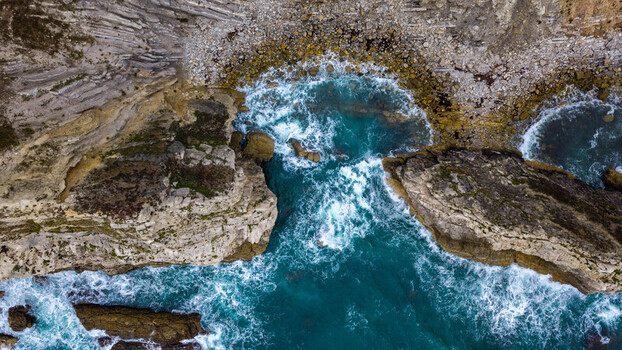 Aerial view of Lulworth Cove with rugged limestone cliffs and turquoise sea water crashing against the rocky coastline in the United Kingdom.