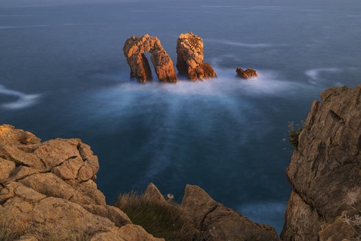 View of the Urro del Manzano sea arch and rugged rock formations surrounded by misty long exposure waves during sunset Liencres Cantabria Spain. View of the Urro del Manzano sea arch and rugged rock formations surrounded by misty long exposure waves during sunset Liencres Cantabria Spain.