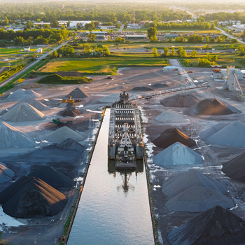 Aerial view of a cargo ship navigating a narrow waterway surrounded by large piles of gravel and industrial aggregate at sunset in Bay City Michigan United States.