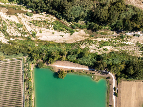 Aerial view of Laghi Santarelli featuring a vibrant turquoise lake surrounded by lush green trees agricultural fields and a winding dirt path in Moresco Marche Italy.