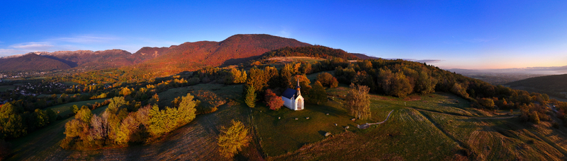 Aerial view of Chapelle Notre Dame de Riantmont surrounded by autumn trees and rolling hills with mountains in the background during sunset Vesancy Auvergne Rhone Alpes France.