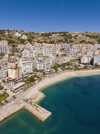 Aerial view of the coastal city of Saranda with dense urban architecture sandy beach and a pier extending into the clear blue waters of the Ionian Sea in Saranda Vlore County Albania. Aerial view of the coastal city of Saranda with dense urban architecture sandy beach and a pier extending into the clear blue waters of the Ionian Sea in Saranda Vlore County Albania.