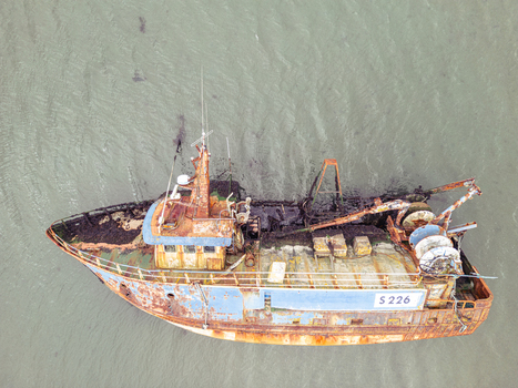 Aerial view of a rusty abandoned fishing boat with registration S 226 resting in shallow murky water with seaweed visible on the deck. Aerial view of a rusty abandoned fishing boat with registration S 226 resting in shallow murky water with seaweed visible on the deck.