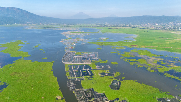 Aerial view of Lake Rawa Pening featuring numerous floating fish farms amidst lush green vegetation and calm waters with distant mountains in Waduk Rawapening Central Java Indonesia. Aerial view of Lake Rawa Pening featuring numerous floating fish farms amidst lush green vegetation and calm waters with distant mountains in Waduk Rawapening Central Java Indonesia.
