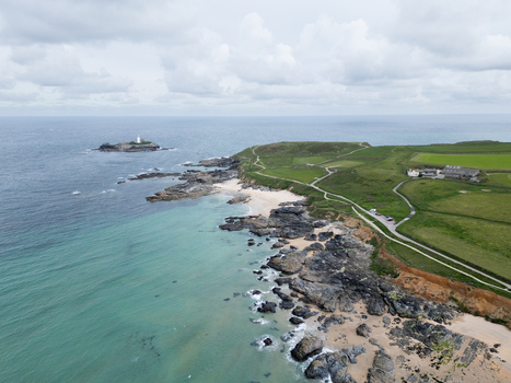 Aerial view of Godrevy Beach featuring the iconic lighthouse on a rocky island turquoise waters and lush green coastal fields under a cloudy sky in Gwithian England United Kingdom. Aerial view of Godrevy Beach featuring the iconic lighthouse on a rocky island turquoise waters and lush green coastal fields under a cloudy sky in Gwithian England United Kingdom.
