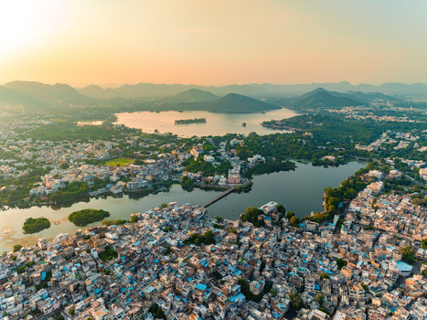 Aerial view of the historic city of Udaipur with its dense urban architecture shimmering lakes and surrounding hills under a warm sunset sky in Udaipur Rajasthan India.