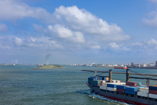 Aerial view of the Port of Rotterdam with a container ship navigating the waterway past a small island with wind turbines under a blue sky in Rotterdam Zuid Holland Netherlands. Aerial view of the Port of Rotterdam with a container ship navigating the waterway past a small island with wind turbines under a blue sky in Rotterdam Zuid Holland Netherlands.