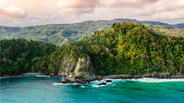 Aerial view of Modangan Beach featuring lush green cliffs rocky outcrops and turquoise waters under a soft sunset sky in Donomulyo East Java Indonesia. Aerial view of Modangan Beach featuring lush green cliffs rocky outcrops and turquoise waters under a soft sunset sky in Donomulyo East Java Indonesia.
