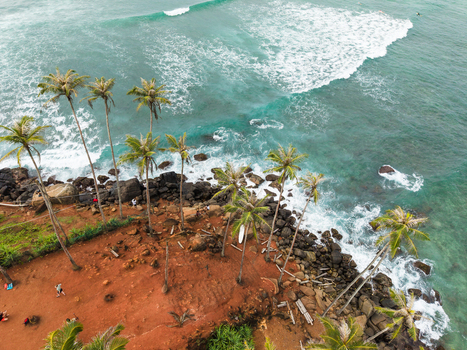 Aerial view of Coconut Tree Hill featuring tall palm trees on a reddish cliff overlooking turquoise ocean waves and rocky shoreline with people in Mirissa Southern Province Sri Lanka. Aerial view of Coconut Tree Hill featuring tall palm trees on a reddish cliff overlooking turquoise ocean waves and rocky shoreline with people in Mirissa Southern Province Sri Lanka.