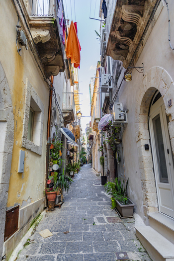 View of a narrow cobblestone street flanked by weathered buildings laundry strung overhead and potted plants adding touches of green Ortigia Sicily Italy.