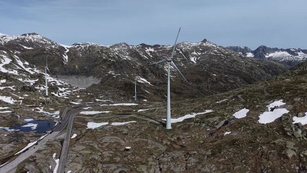 Aerial view of San Gottardo Wind Park featuring wind turbines among snowy mountain peaks and a winding road in the Gotthard Pass Airolo Ticino Switzerland.