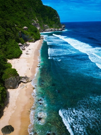 Aerial view of Pantai Nunggalan featuring a pristine sandy beach turquoise ocean waves crashing against the shore and lush green cliffs in Kecamatan Kuta Selatan Bali Indonesia.