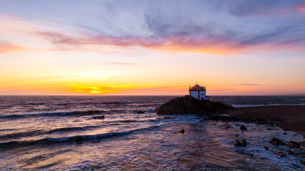 Aerial view of Capela do Senhor da Pedra on a rocky outcrop with waves crashing and a vibrant sunset sky Miramar Porto Portugal.