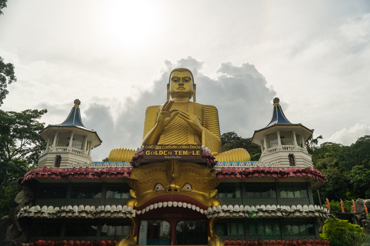 View of the giant golden Buddha statue at the Dambulla Golden Temple surrounded by lush green trees under a cloudy sky Dambulla Central Province Sri Lanka.