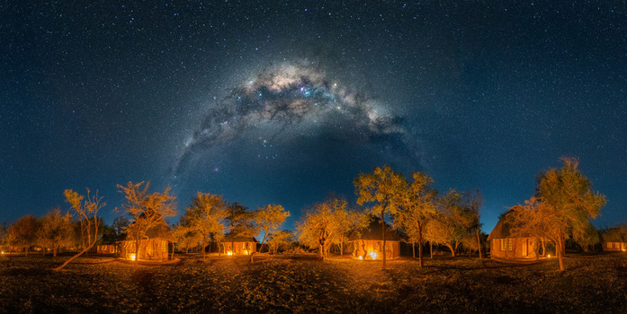 View of Ndlovu Camp in Hlane Royal National Park featuring illuminated traditional huts under a vibrant Milky Way arch and starry night sky in Simunye Lubombo Region Eswatini.
