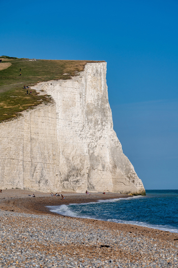 View of Seven Sisters Cliffs with towering white chalk cliffs a pebble beach with people and the calm blue sea under a clear sky in Exceat England United Kingdom. View of Seven Sisters Cliffs with towering white chalk cliffs a pebble beach with people and the calm blue sea under a clear sky in Exceat England United Kingdom.