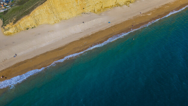 Aerial view of the sandy beach and golden sandstone cliffs at West Bay with turquoise sea water and people walking along the shore in West Bay England United Kingdom. Aerial view of the sandy beach and golden sandstone cliffs at West Bay with turquoise sea water and people walking along the shore in West Bay England United Kingdom.
