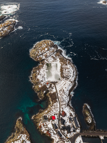 Aerial view of a snow dusted island village with vibrant red roofs contrasting against the dark swirling sea Henningsv&aelig;r Nordland Norway.